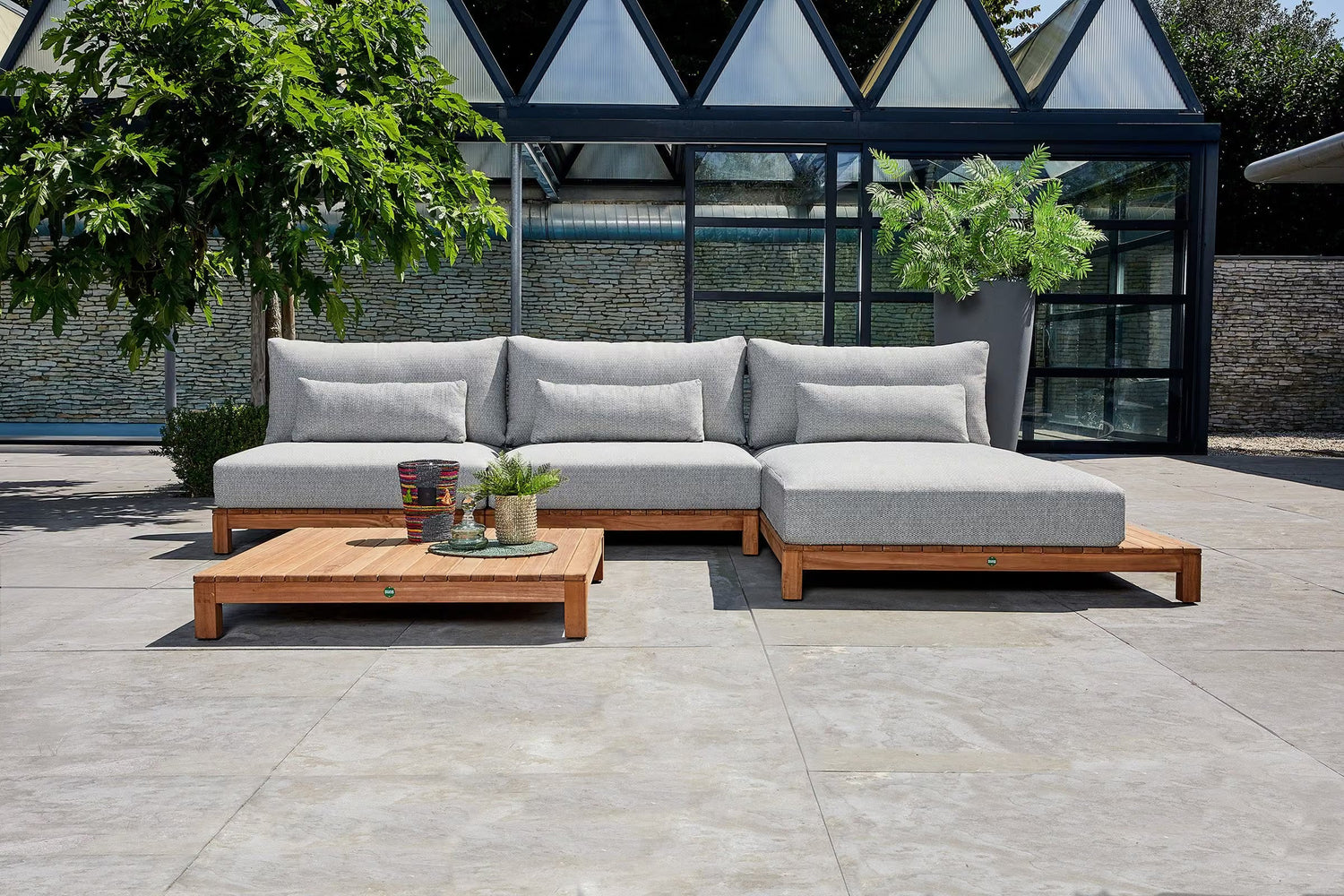 Outdoor sectional sofa with light gray cushions and a wooden base, arranged on a paved patio in front of a modern glass conservatory. A matching wooden coffee table sits in front of the sofa.