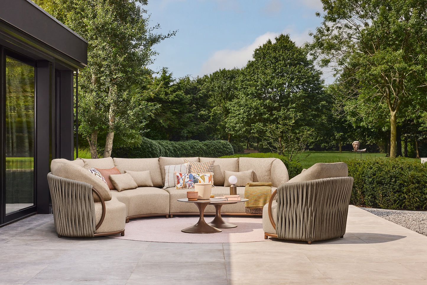 Outdoor patio with beige sectional sofa, chairs, and coffee table in a garden setting.