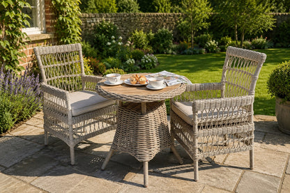 A three-piece outdoor wicker furniture set consisting of a round woven table and two matching armchairs with cream cushions, set on a stone patio in a lush garden.