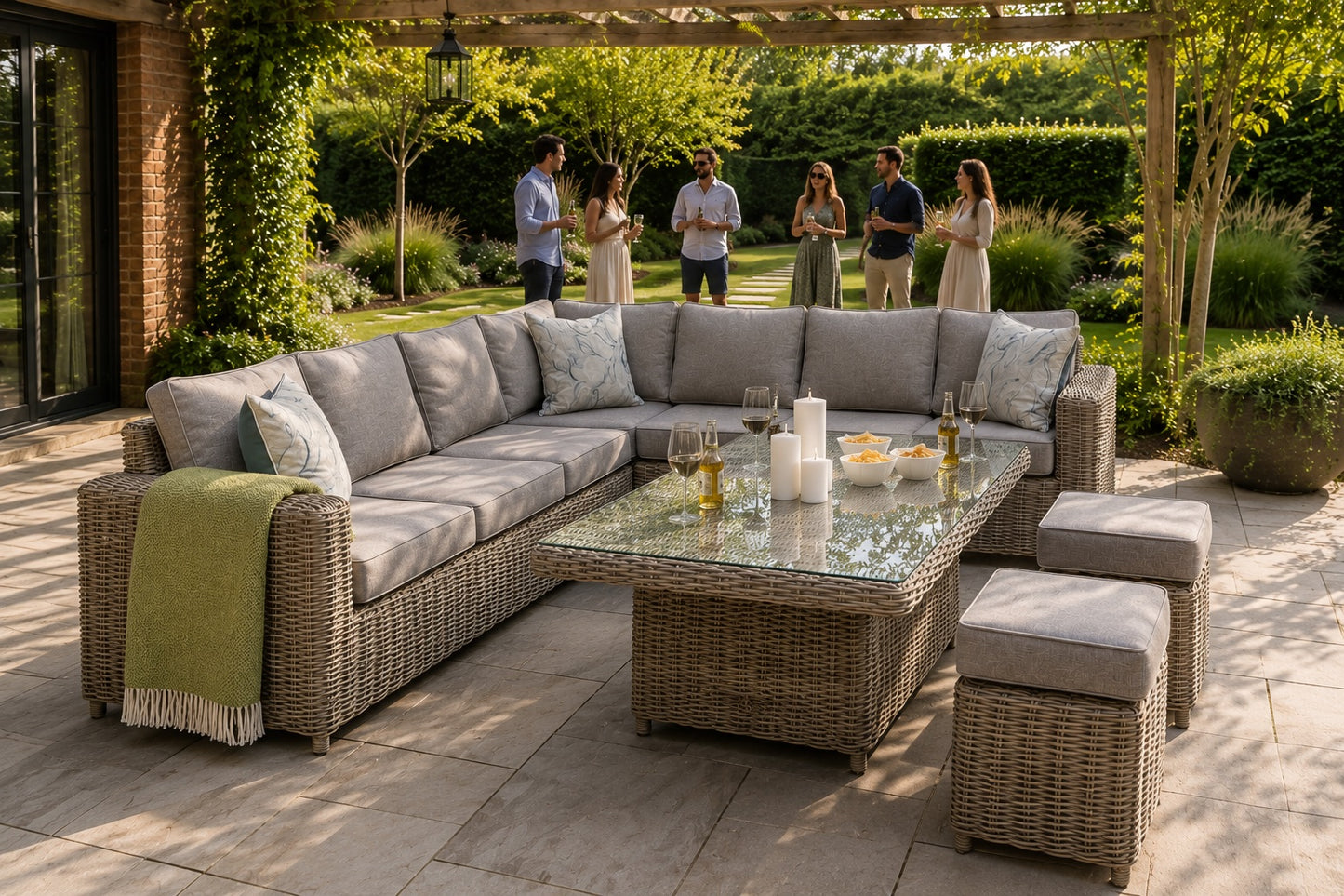 An outdoor patio conversation set with a wicker sectional sofa, matching ottomans, and a glass-top coffee table, staged on a stone patio under a pergola with garden views and people in the background.