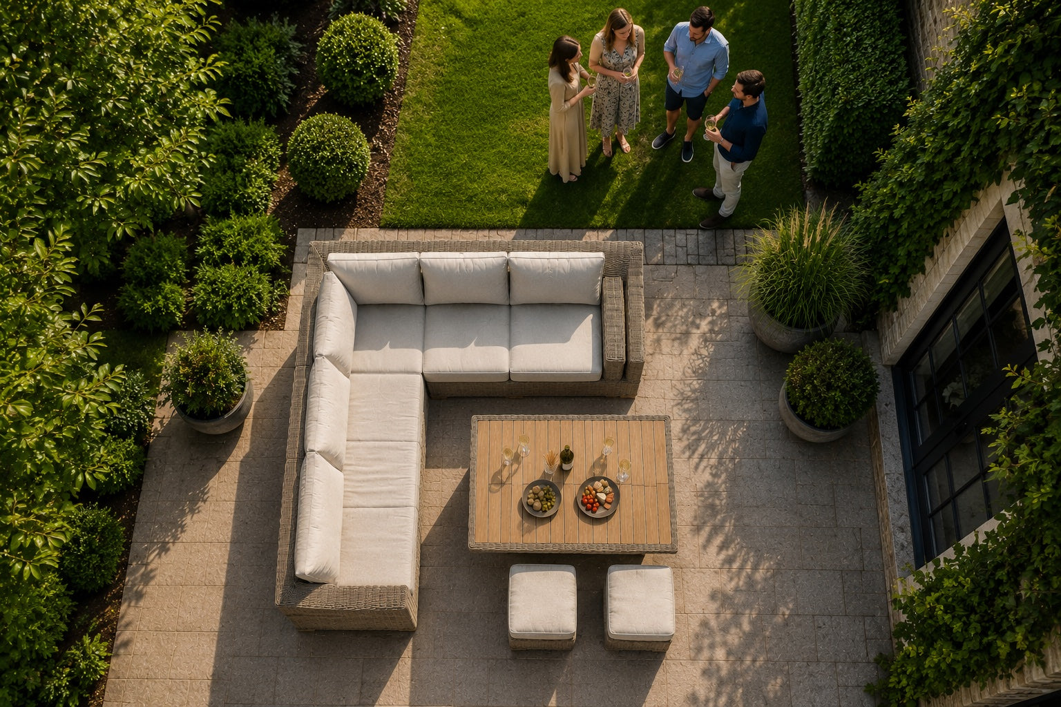 Overhead view of an outdoor patio furniture set on a stone patio, including a white sectional sofa, a wooden coffee table, two matching ottomans, and a grassy garden area with three people standing nearby.