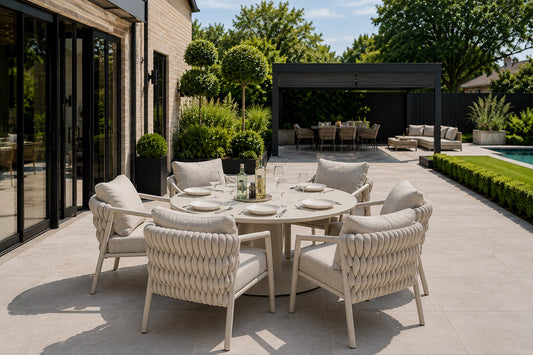 An outdoor dining set with a round woven table and four matching armchairs in a modern backyard patio setting. The furniture is upholstered in light beige fabric with cream-colored frames, arranged on a light stone patio with a house, greenery, and a pool visible in the background.