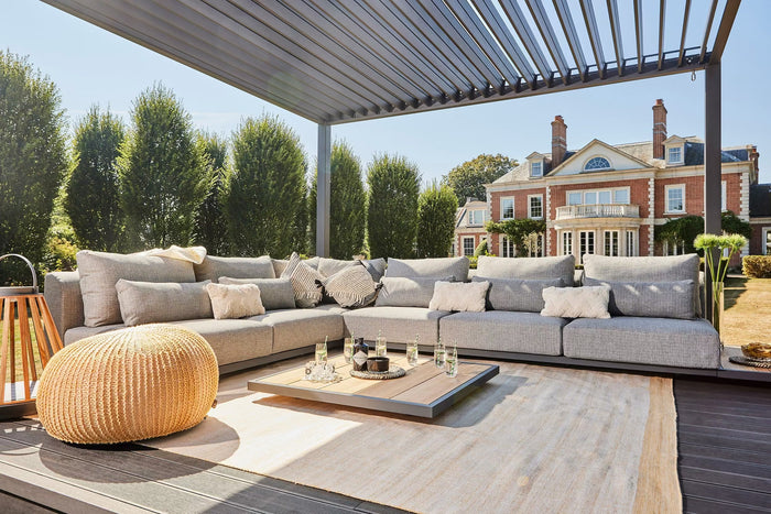 Outdoor patio furniture set under a slatted roof pergola, featuring a light gray modular sectional sofa, a woven natural-fiber pouf, a low wooden coffee table, and a light rug, with a large garden house and trees in the background.