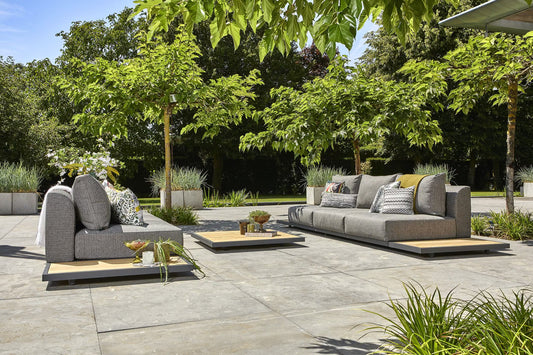 Modern outdoor patio furniture set with a gray modular sofa, matching lounge chair, and two low wooden coffee tables on a stone patio, surrounded by lush green trees and plants.