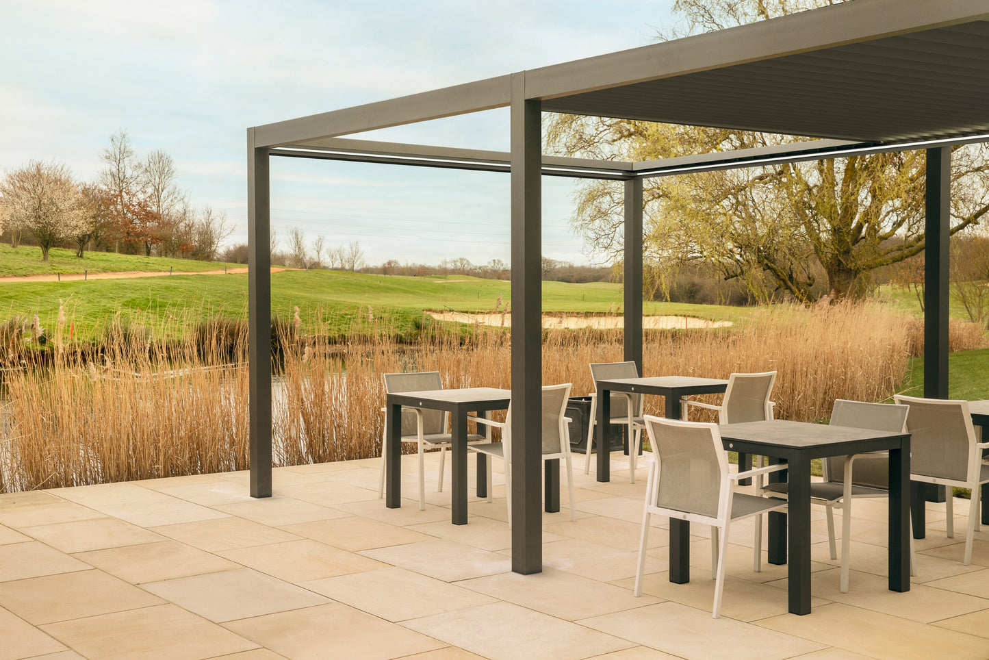Outdoor patio with tables and chairs under a pergola, surrounded by nature.