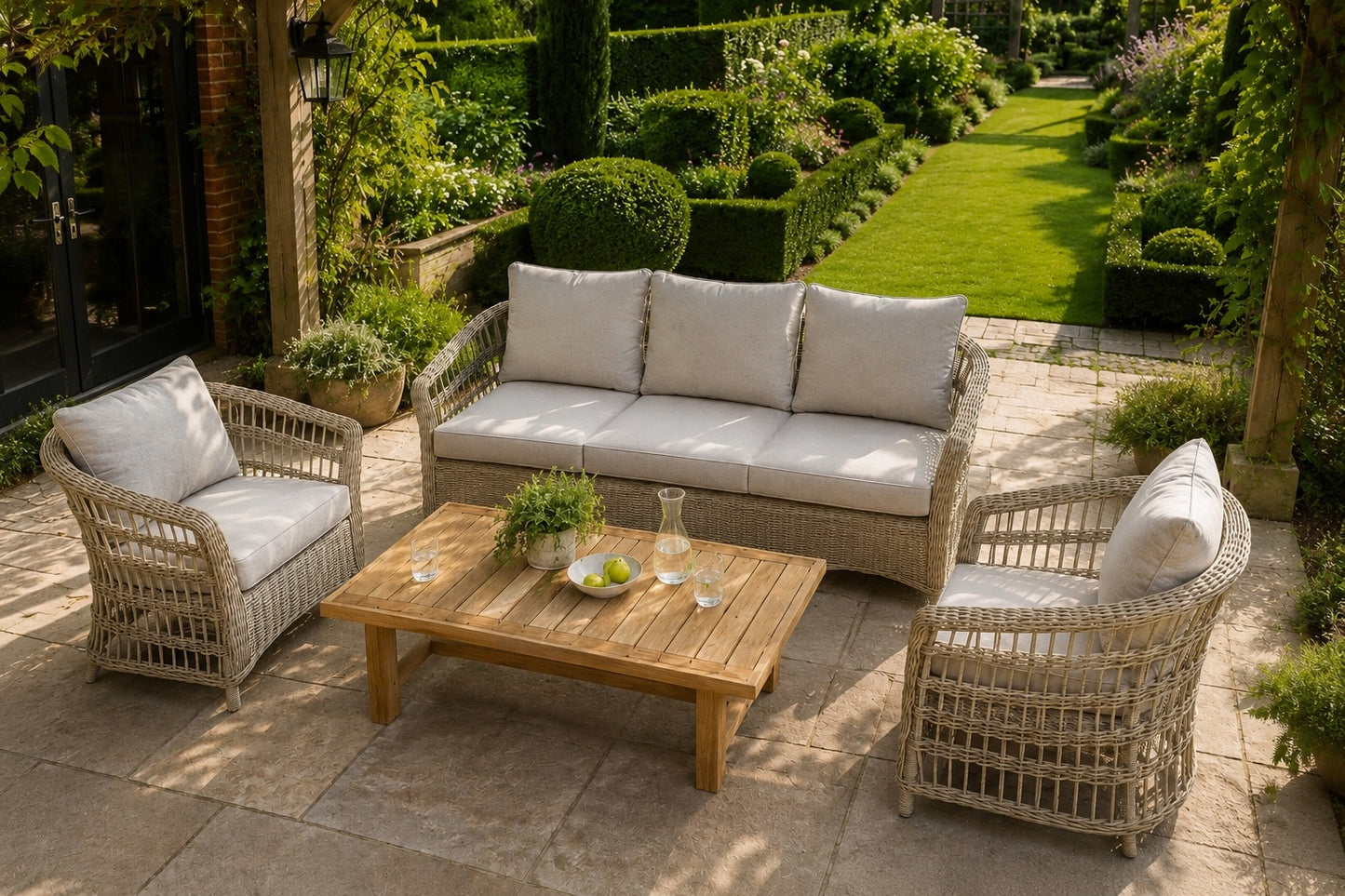 A 4-piece outdoor patio furniture set including a 3-seater sofa, 2 armchairs, and a rectangular coffee table, made of light beige woven wicker with off-white cushions, arranged on a stone patio in a lush garden setting.