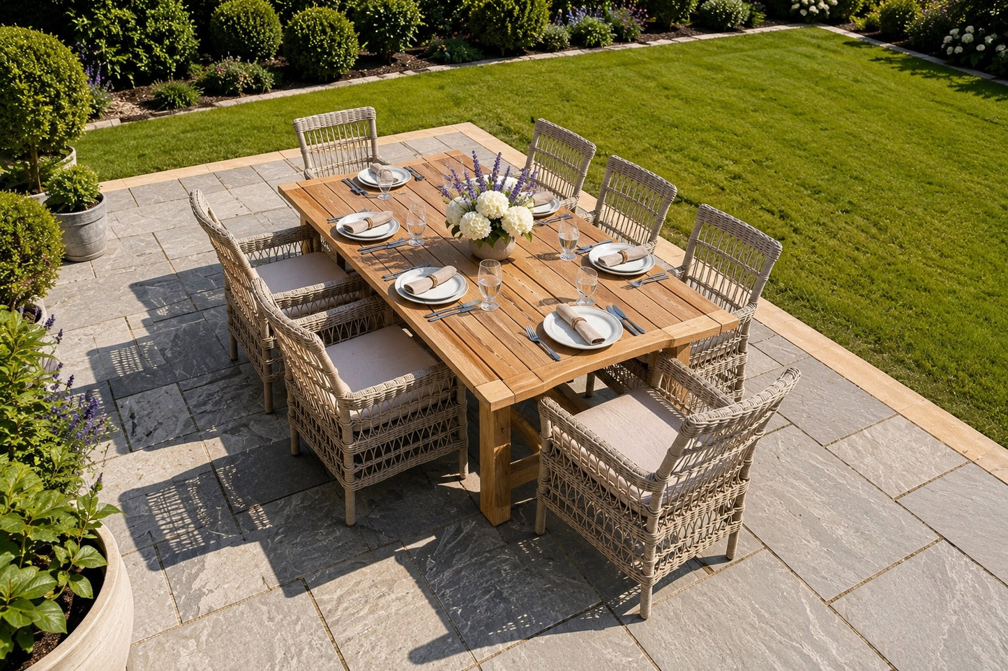 An outdoor dining set on a stone patio in a landscaped garden, featuring a rectangular wooden dining table surrounded by eight woven wicker chairs with beige cushions.