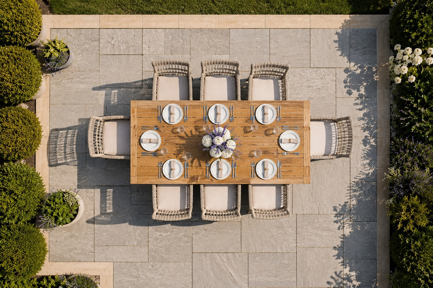 Overhead view of an outdoor dining set on a stone patio, featuring a rectangular wooden dining table surrounded by eight woven wicker chairs with cream cushions, set with white plates and a floral centerpiece.