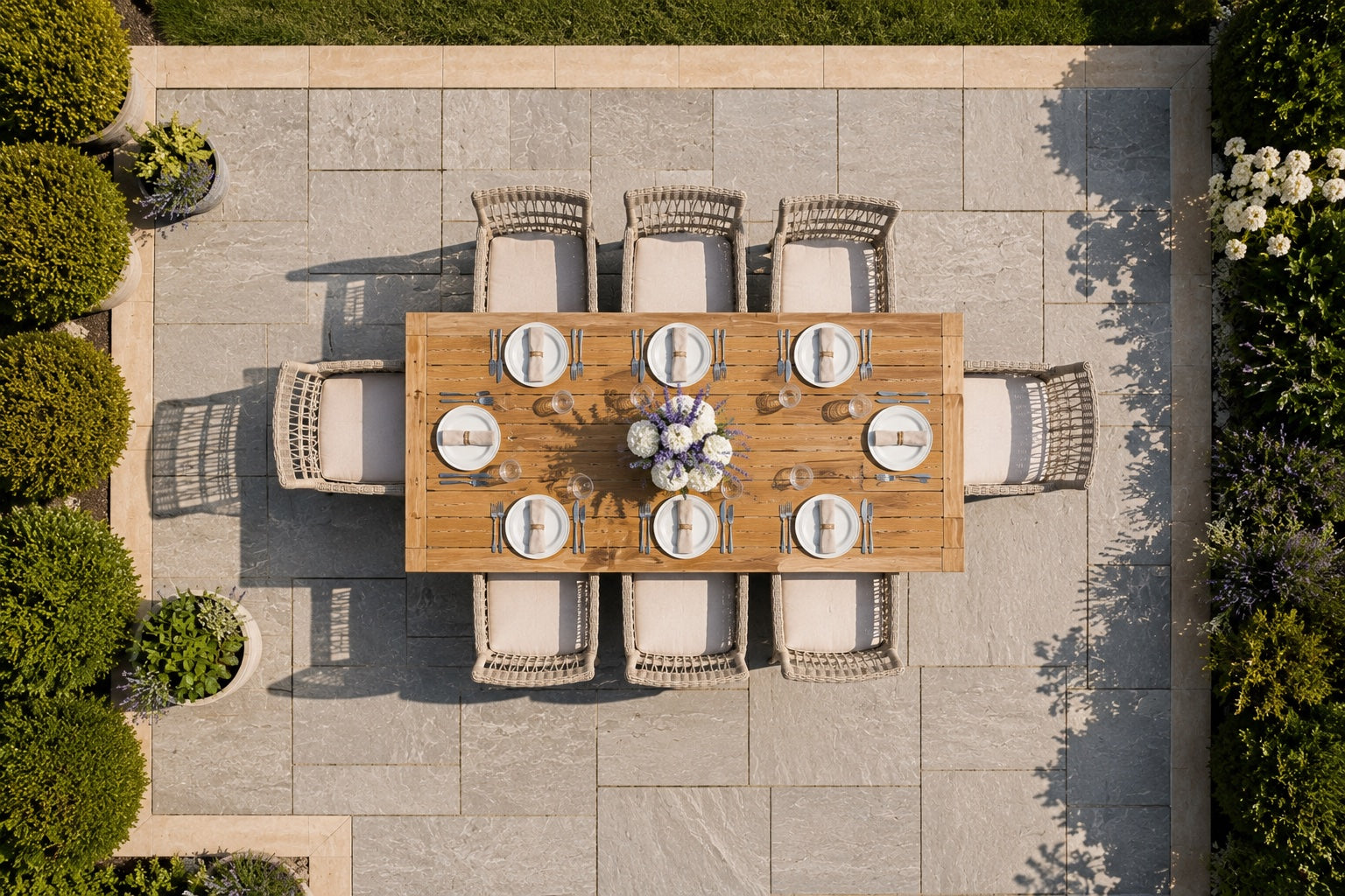 Overhead view of an outdoor dining set on a stone patio, featuring a rectangular wooden dining table surrounded by eight woven wicker chairs with cream cushions, set with white plates and a floral centerpiece.