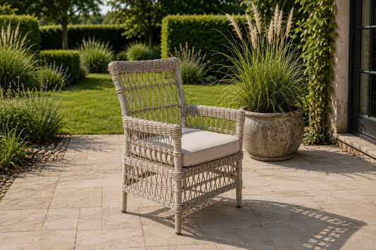A light beige woven rattan armchair with a cream-colored seat cushion, positioned on a stone patio in a beautifully landscaped garden setting.