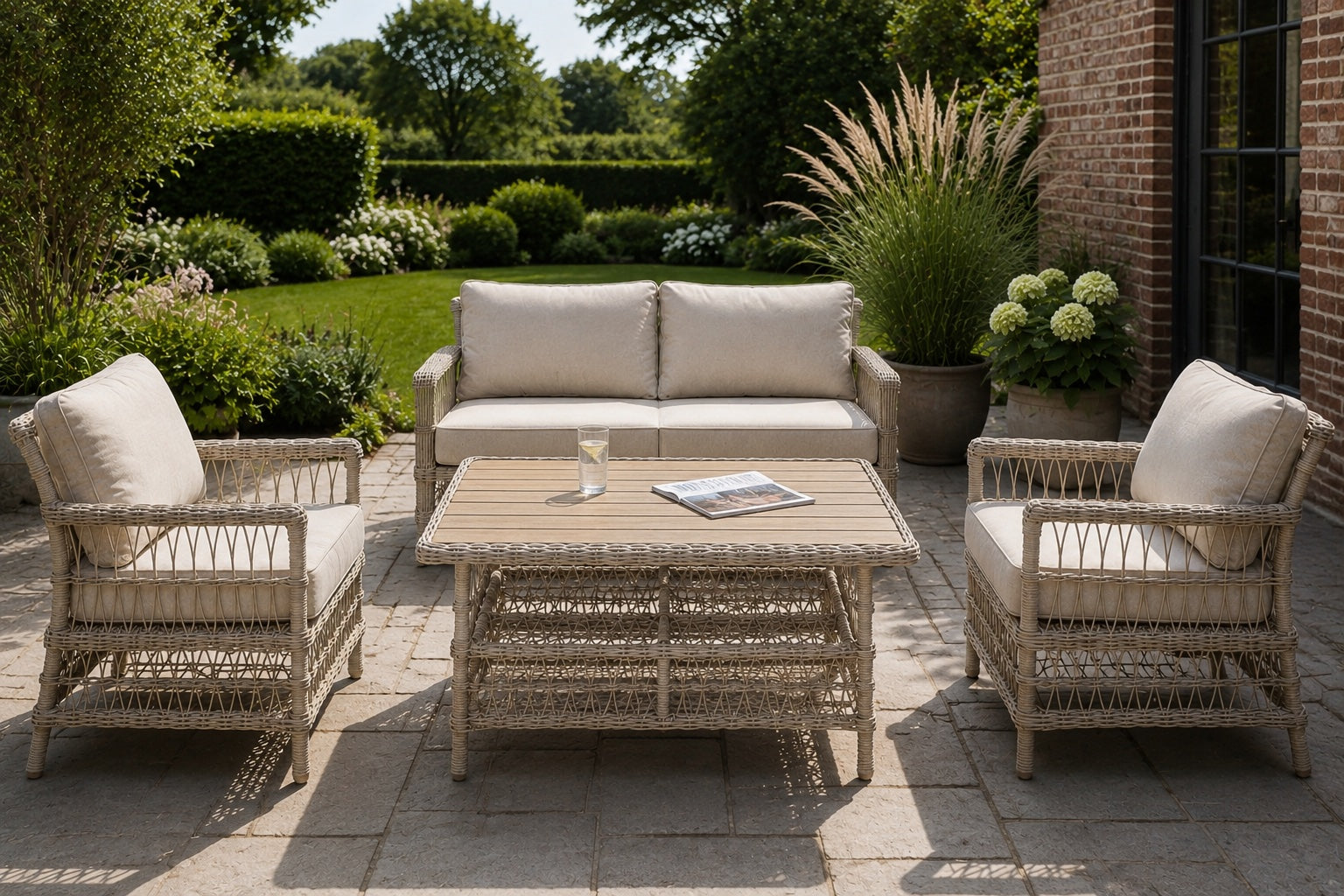 A 4-piece outdoor patio furniture set including a two-seater sofa, two armchairs, and a rectangular coffee table, all in a light natural wicker weave with beige cushions, arranged on a stone patio in a garden setting.