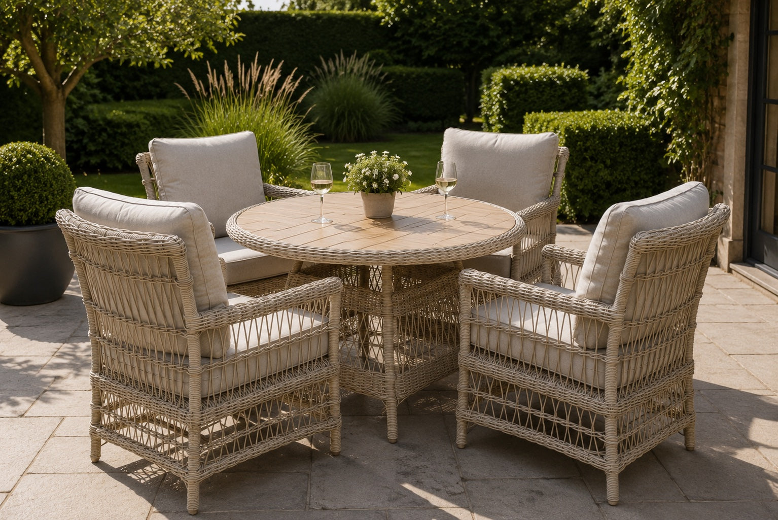 A 4-piece outdoor patio dining set featuring a round wicker table and four matching armchairs with light beige cushions, set on a stone patio in a lush garden.