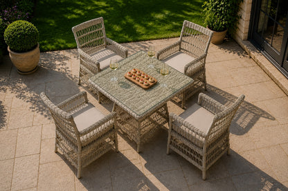 A 5-piece outdoor patio dining set made of woven wicker/rattan, featuring a square table with a green-tinted glass top and four matching armchairs with light beige cushions, set on a stone patio in a garden.
