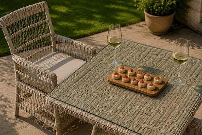 Outdoor patio scene with a woven wicker armchair and matching square table. The table has a glass top and holds a wooden board with small appetizers, plus two wine glasses filled with white wine.