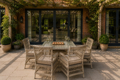 A 5-piece outdoor dining set consisting of a square glass-top table and four woven wicker armchairs, arranged on a stone patio in front of a brick house with black-framed glass doors.