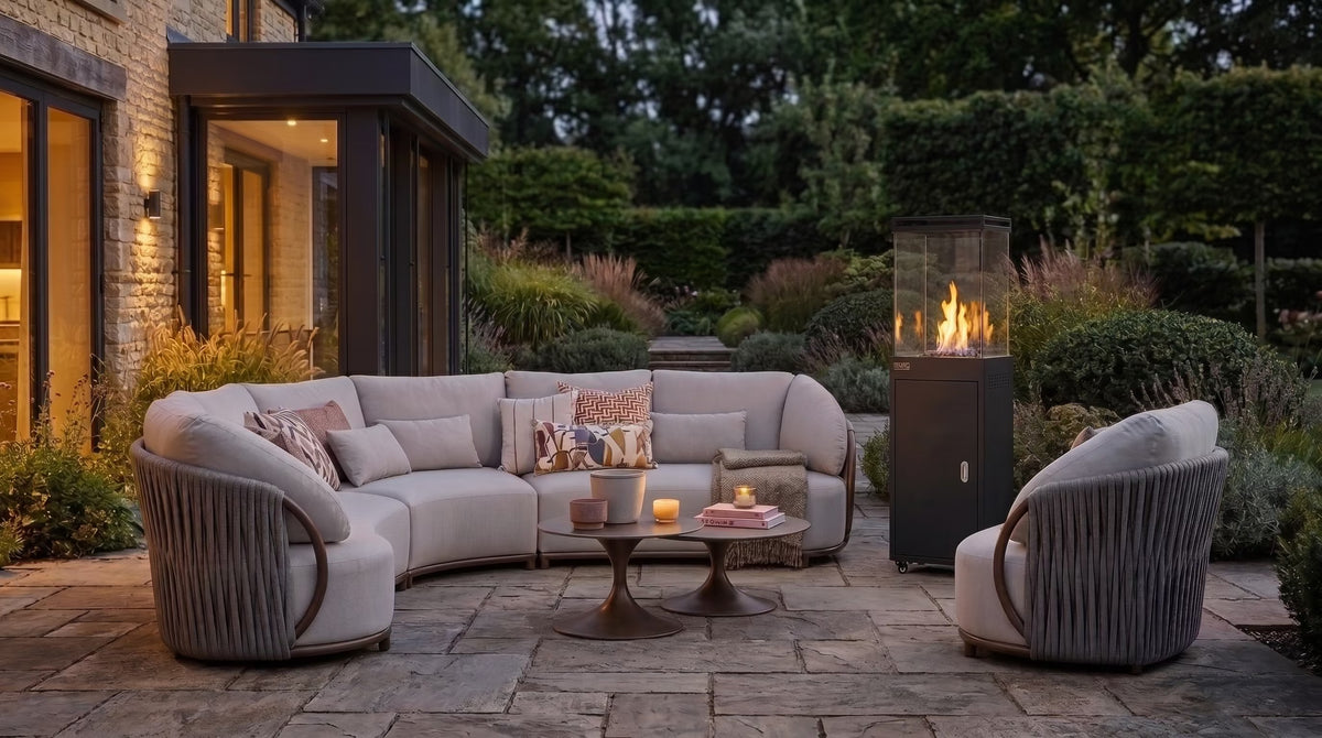 Outdoor patio furniture set including a curved sectional sofa, two matching armchairs, a round coffee table, and a tall black outdoor fire pit, arranged on a stone patio in a landscaped garden at dusk.