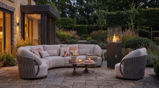 Outdoor patio furniture set including a curved sectional sofa, two matching armchairs, a round coffee table, and a tall black outdoor fire pit, arranged on a stone patio in a landscaped garden at dusk.